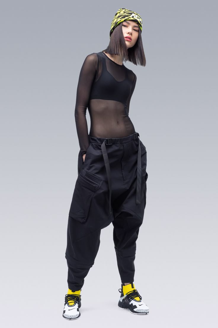 a woman in streetwear fashion. she is wearing a black mesh top over a black bra top, baggy black cargo pants with a black belt, a yellow and black hat, yellow socks, and white sneakers with black and yellow accents. she is standing in a studio with a gray background, posing for the camera.