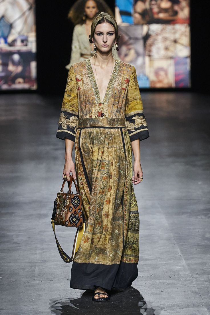 a female model walks the runway wearing a long floral print dress, black sandals, and a small brown handbag with a long strap. she has a headband in her hair and is wearing earrings. the background is blurred with other models and the audience.