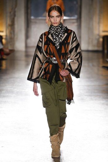 a fashion model walks the runway wearing a stylish outfit.  the outfit consists of a black and beige geometric patterned poncho, olive green cargo pants, tan suede boots, and several accessories including a brown leather bag, a black and white patterned scarf, and a wide belt with a large buckle.  the model's hair is styled up, and her overall look is bohemian and westerninspired, fitting for an autumn or winter season. the background suggests a runway setting, perhaps in a large hall or building.