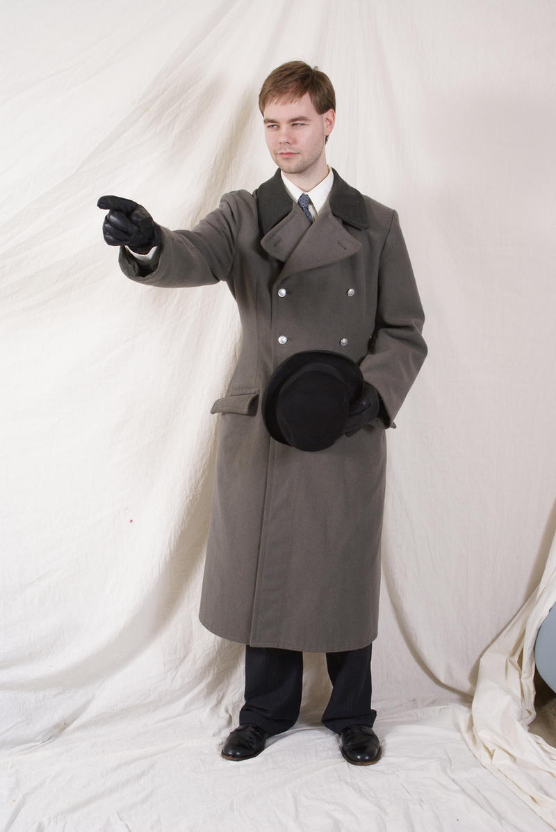 a man dressed in a long gray coat, black dress pants, and black dress shoes stands against a white backdrop. he wears black gloves and holds a black hat in his left hand. he points with his right hand.