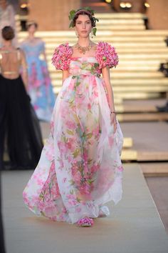 a woman wearing a beautiful white and pink floral dress with a sheer overlay and floral details on the shoulders and sleeves. she is walking down a runway and smiling as she poses for the cameras. her hair is pulled back with a floral crown.