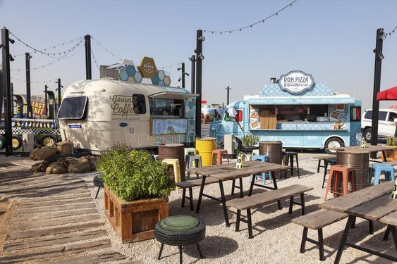 a picture of a food truck scene. it is an outdoor eating area with various tables and chairs. the image shows two food trucks: one is a gourmet coffee truck and the other is a don pizza truck. both trucks have large signs with their names on them, in addition to other details about their offerings. the scene is set up with picnic tables, chairs, and stools for customers to enjoy their food. the scene has multiple plants and bushes throughout the scene.
