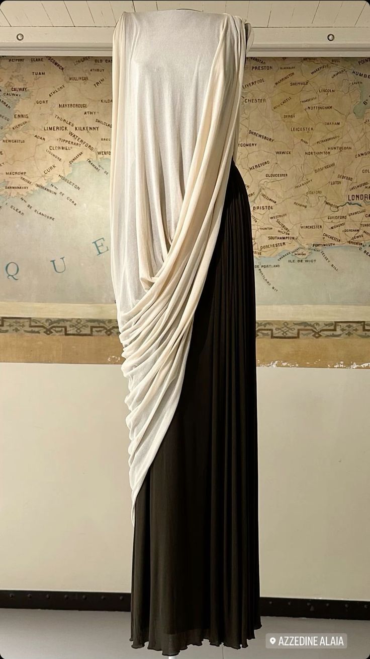a long dress on a mannequin. the dress has a black pleated skirt and a creamcolored draped top. a map is behind the mannequin. the dress is labeled "azzedine alaia."