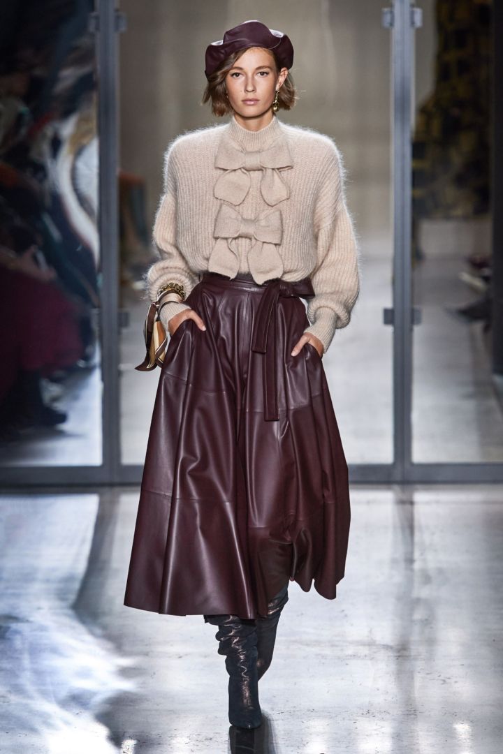 full shot of a fashion model showcasing a fall/winter outfit on a runway. the model is wearing a beige oversized knit sweater with bow details, a burgundy leather midi skirt,  burgundy high boots, and a burgundy beret. she carries a small bag in one hand.  the background is a minimalist grey and silver setting typical of a fashion show. the overall style is sophisticated and elegant, reflecting current trends in autumn/winter fashion.