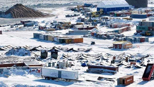 a wintery construction site with a large pile of snow and several shipping containers scattered throughout. there is a large white tent in the background and several pieces of heavy equipment. the scene is cold and desolate.