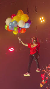 a man in a red shirt is singing into a microphone on stage at a concert. he is surrounded by colorful balloons floating above him. he is smiling and appears to be enjoying himself.  the stage is lit by spotlights.