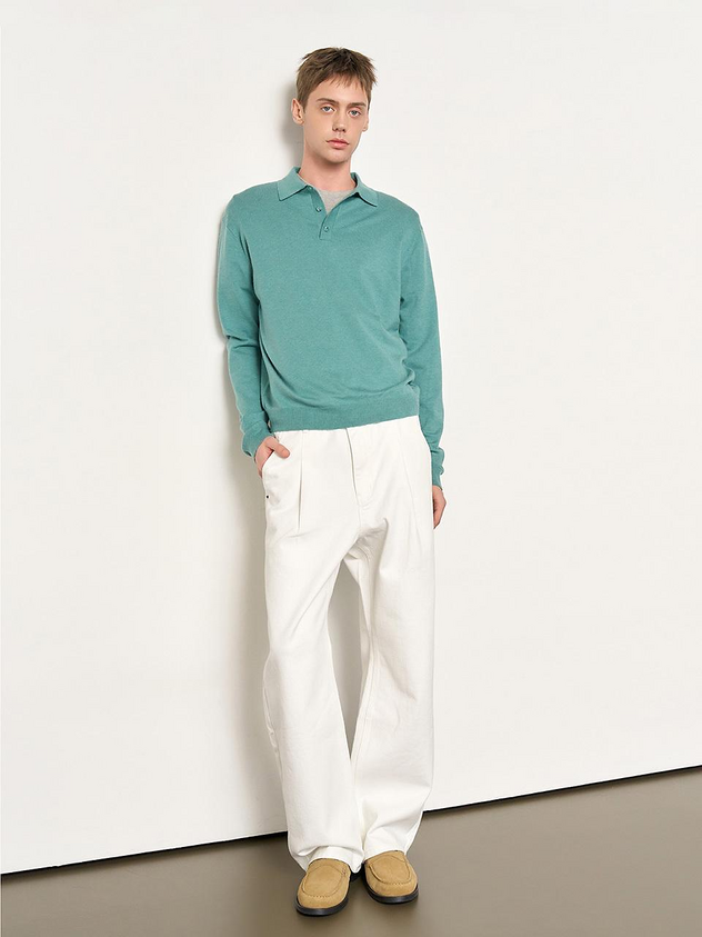 a man wearing a teal polo sweater and white pants, with tan shoes, stands against a white wall.