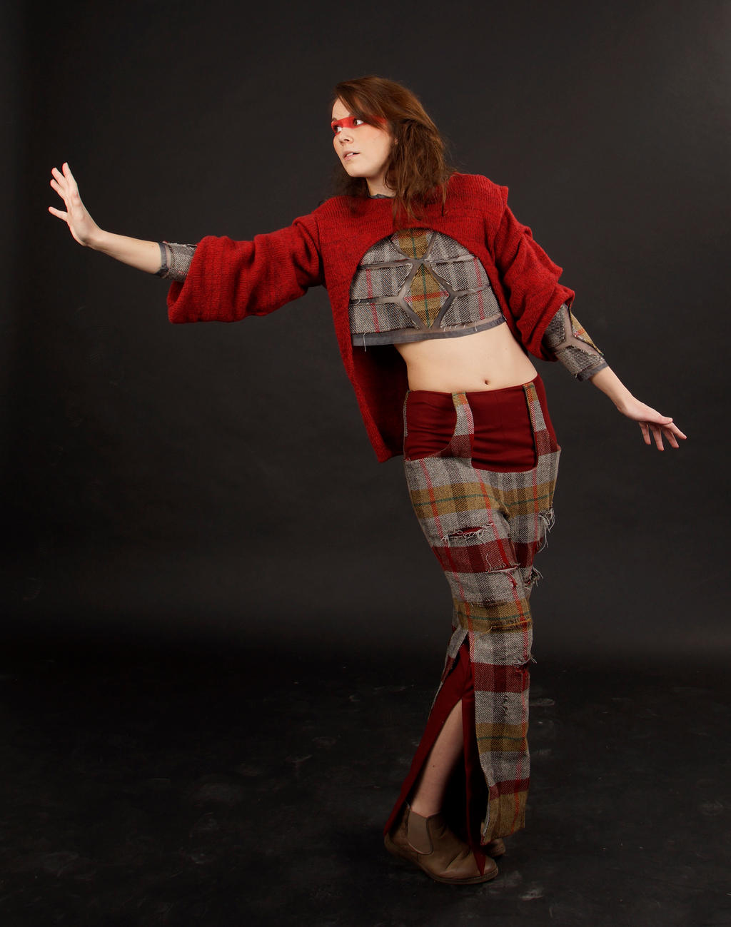 a full shot of a female model, showcasing a unique outfit.  she is wearing a red knitted sweater with a cropped, patterned underlayer and plaid pants with distressed detailing. the pants feature a split leg design.  she has red accents on her face and is posing dynamically with one arm outstretched. the background is a plain, dark backdrop. the overall image suggests a high fashion or artistic fashion photoshoot.