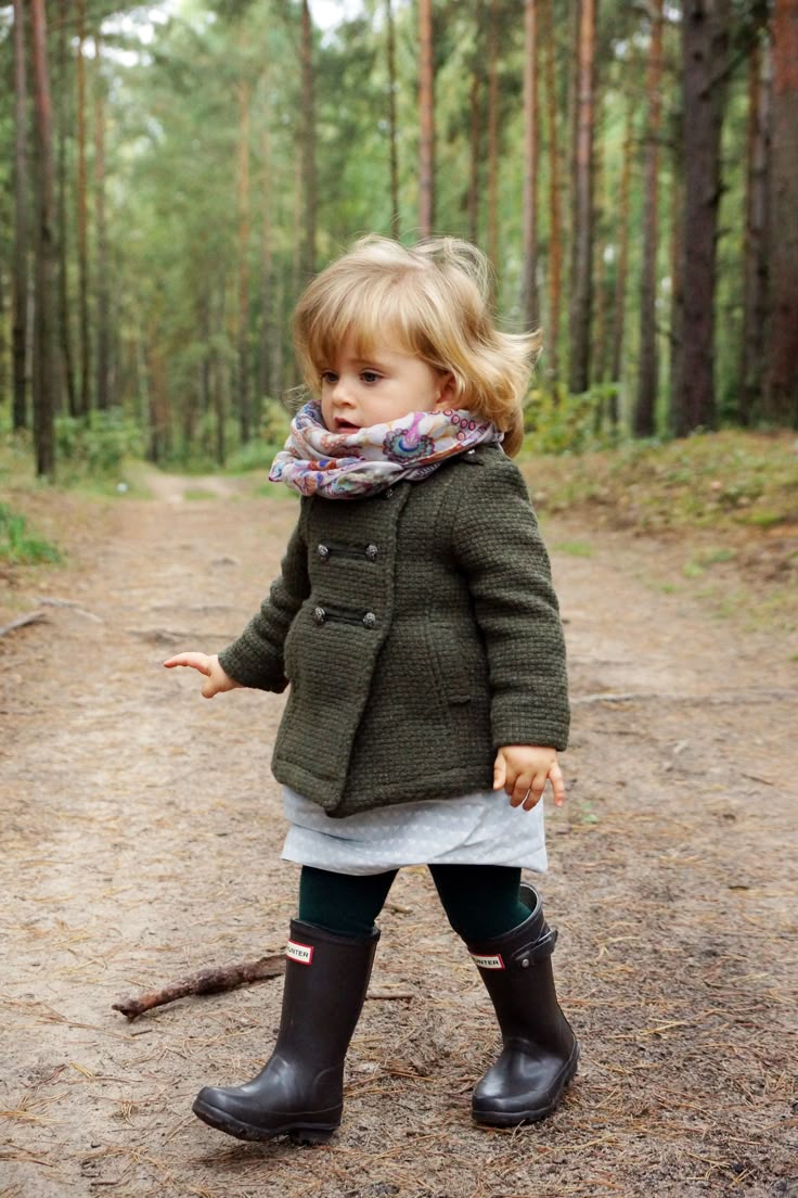 a young toddler girl is walking on a dirt path in the woods. she is wearing a dark green wool coat, a lightcolored scarf with a floral pattern, a light gray dress, dark green tights, and dark brown hunter brand rain boots. the background shows tall trees typical of a forest or wooded area. the scene suggests it is autumn, given the clothing and the light.