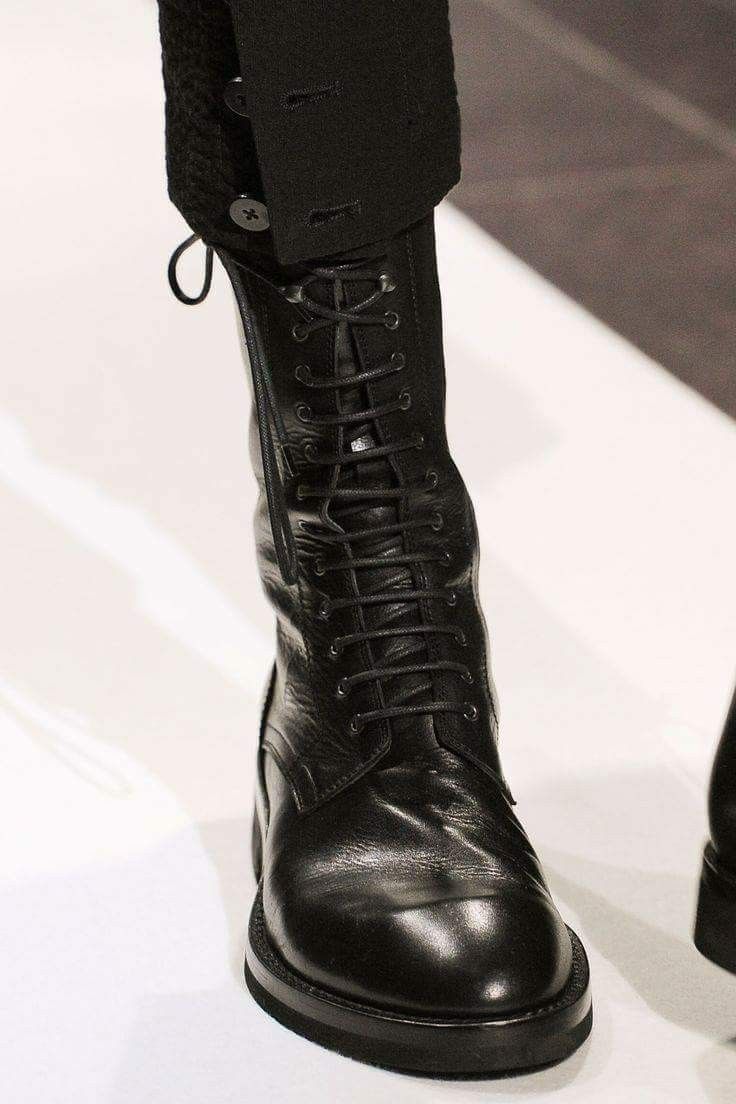 a closeup shot of a black leather laceup boot. the boot is tall and reaches up the calf. the laces are black and are tightly tied. the leather is shiny. the boot is sitting on a white surface. the boot is partially covered by a black garment with button detail.