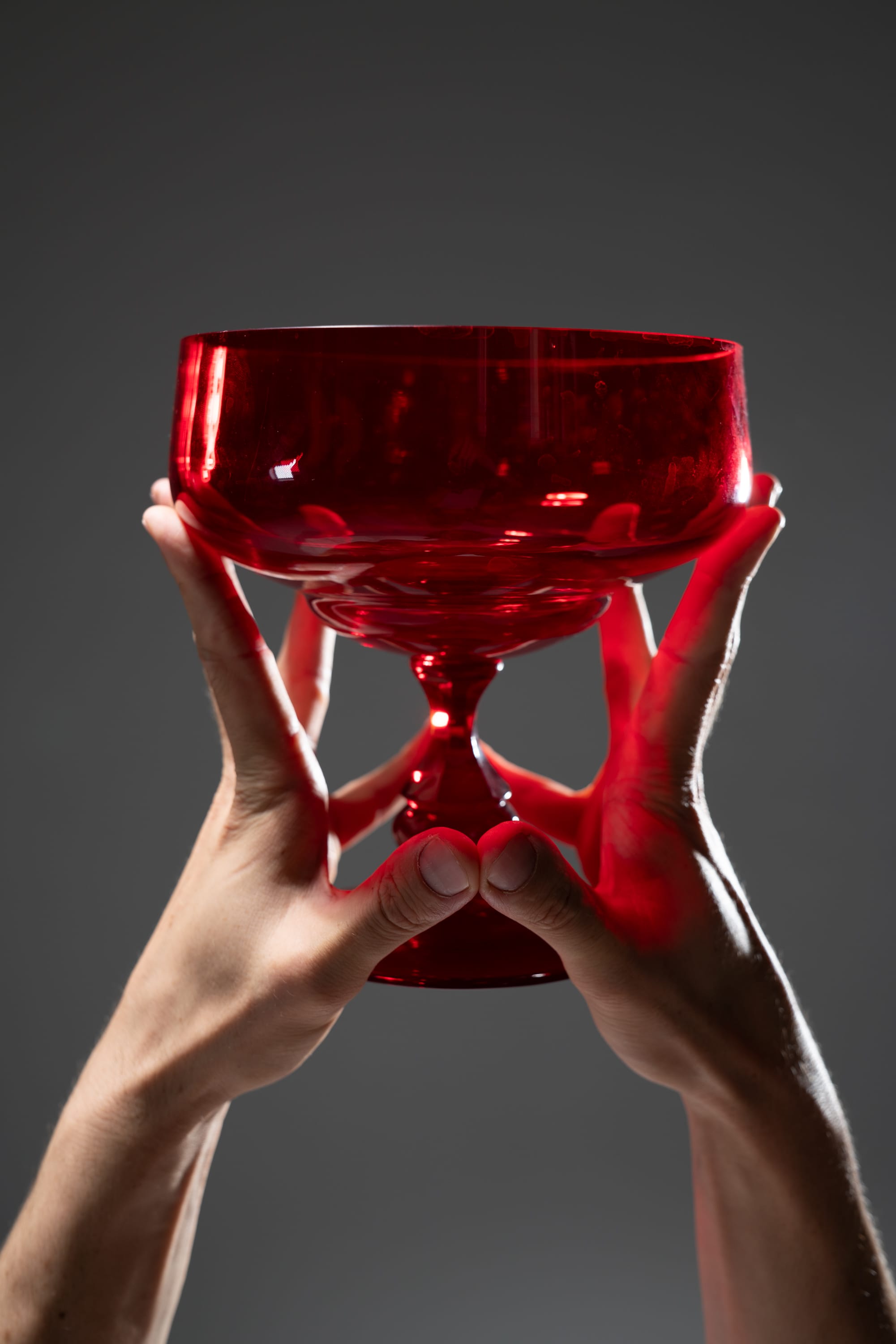 a pair of hands lift a red chalice against a grey background. the bottom of the chalice is brightly lit.