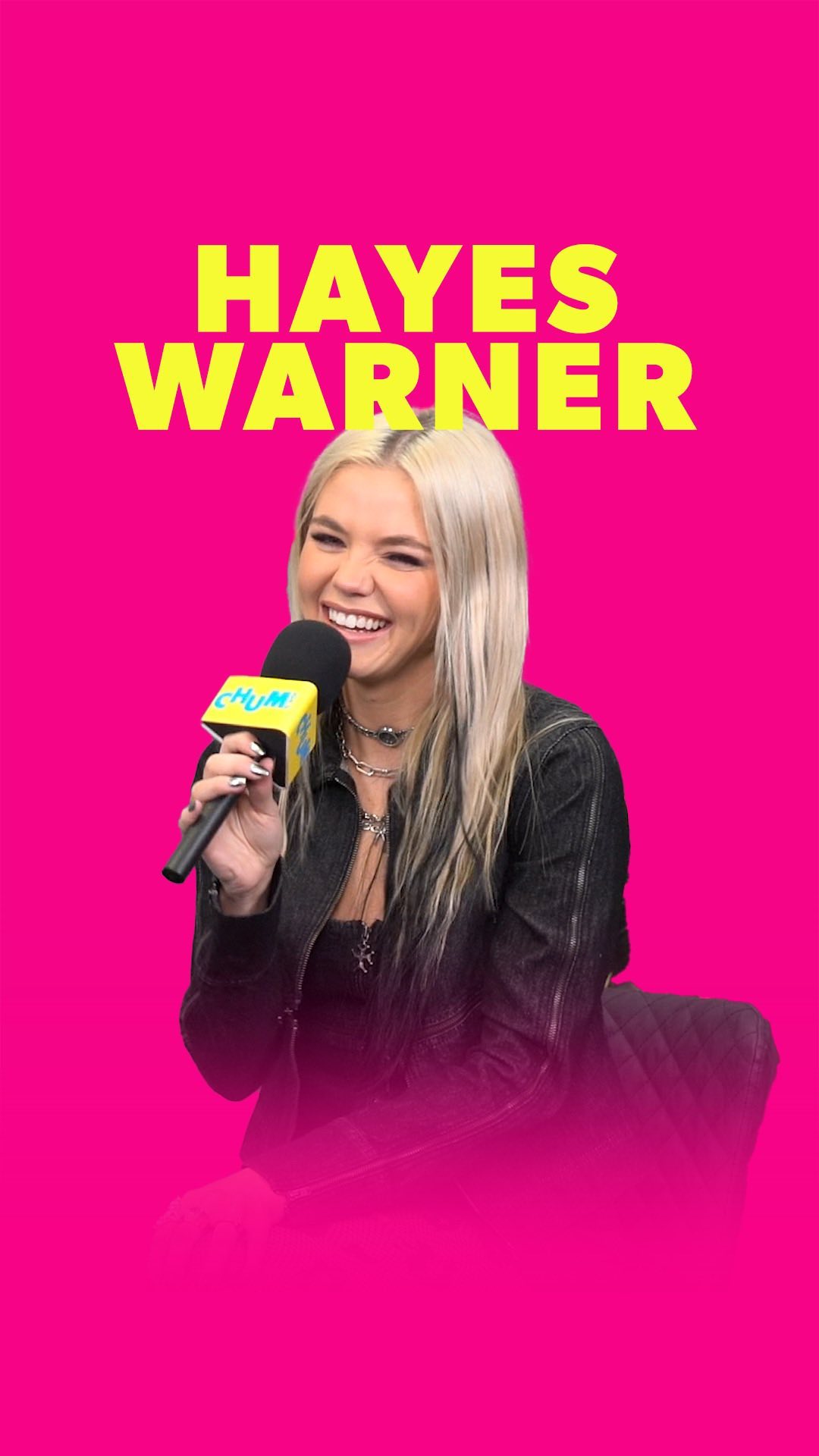 a woman named hayes warner smiles while holding a microphone with the chum logo on a pink background. she is wearing a denim jacket and necklaces.