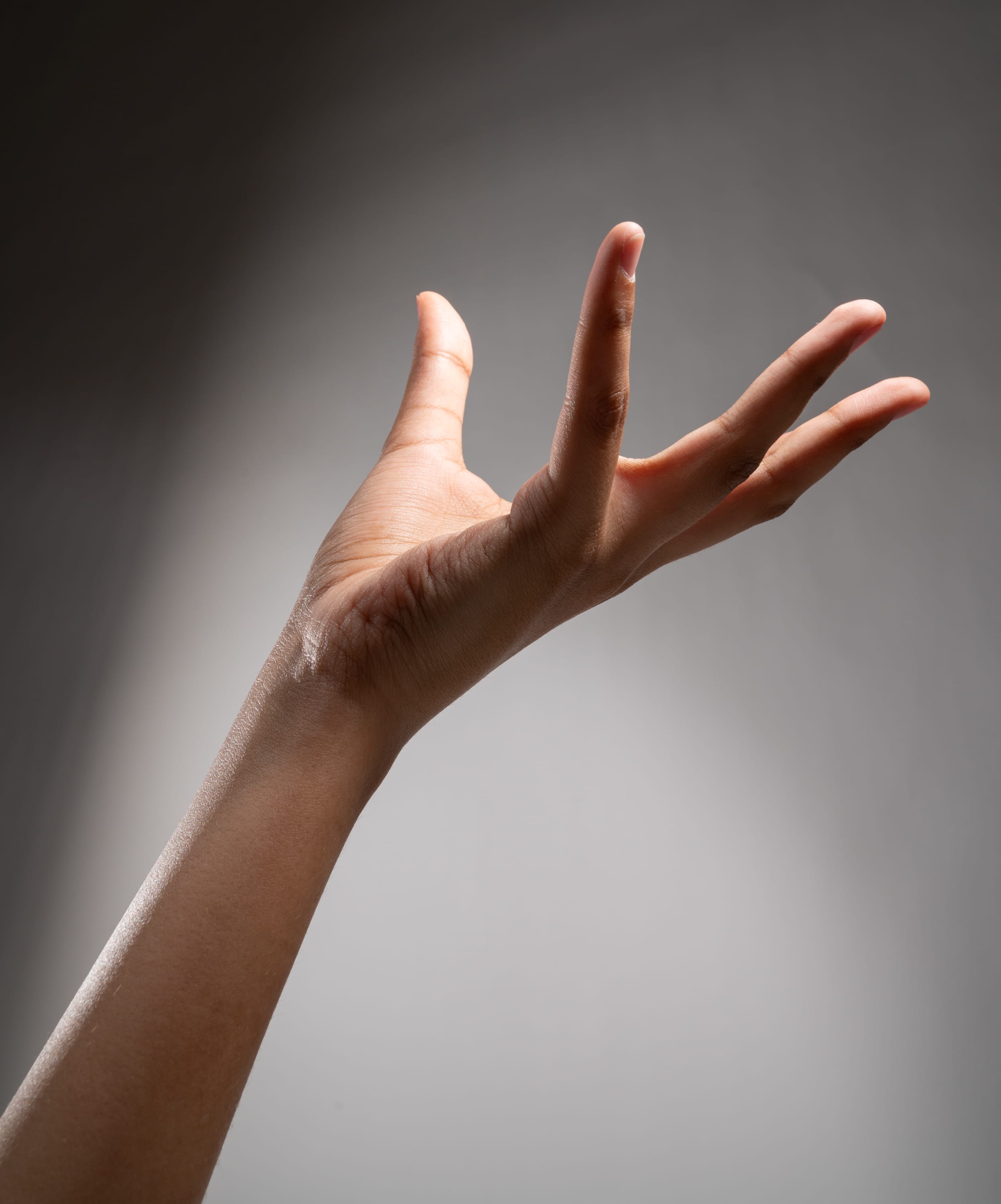 he image shows a single hand reaching upwards against a gray background. the hand is positioned in profile, with the fingers spread slightly apart. the lighting is dramatic, creating a spotlight effect on the hand and highlighting the contours of the skin. the image has a grayscale palette, emphasizing the shape and form of the hand.