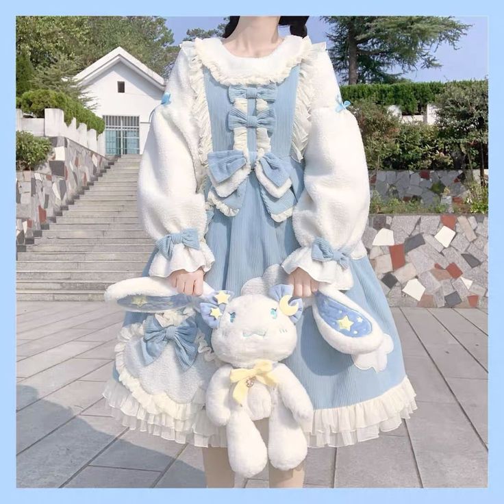 a woman is standing outside on stone stairs while modeling a blue and white lolita fashion dress. she is also holding a white bunny plush. the dress has a peter pan collar, long sleeves with white faux fur, a blue corduroy bodice and skirt, and lace and ribbon embellishments. she is wearing the dress with a white longsleeved shirt underneath.