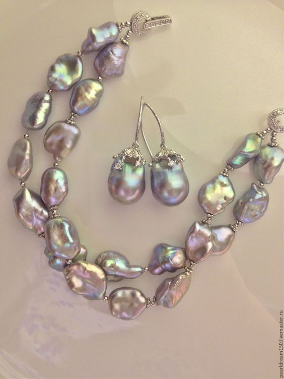 a matching bracelet and earring set made from large, baroque, iridescent keshi pearls set in silver. the pearls range in color from silver to blue to green. the bracelet has two strands of pearls and the earrings are simple drop earrings. the jewelry is lying on a white plate.