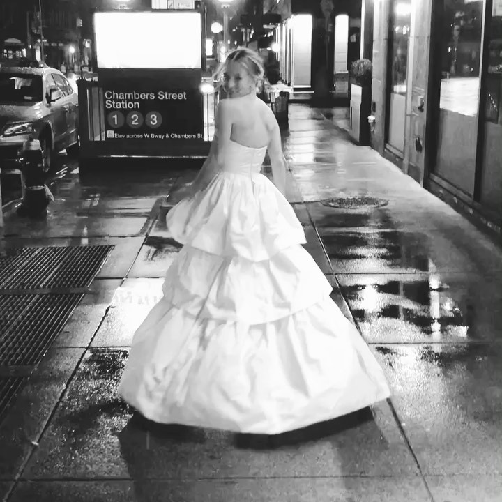 a black and white fulllength shot of a woman in a voluminous, tiered wedding dress walking on a wet city street at night. the background shows the chambers street station sign in new york city.  the woman's back is to the camera, and she appears to be alone. the overall mood is romantic and slightly melancholic.