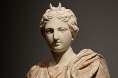 the image is a closeup of a marble bust of a female figure. the figure is wearing a crescent moon on her head and a draped garment. the statue is lit from the front and slightly above, casting shadows on the figure's face and neck. the background is a plain, dark gray wall. the image has a shallow depth of field, focusing on the figure's face.