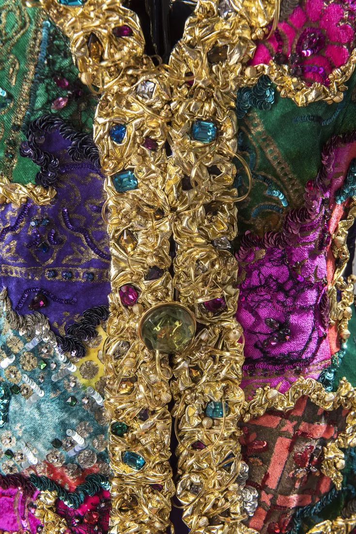closeup detail shot of a richly embellished garment, possibly a jacket. the piece is a patchwork of various fabrics in vibrant colors including purple, green, pink, and teal velvets.  the fabrics are heavily adorned with gold embroidery, sequins, and numerous jewels of varying colors and sizes, including what look like crystals and gemstones. the gold work is highly textured and sculpted, creating a threedimensional effect. the overall impression is one of extreme luxury and ornate detail. the image showcases the intricate craftsmanship and the opulent nature of the garment.