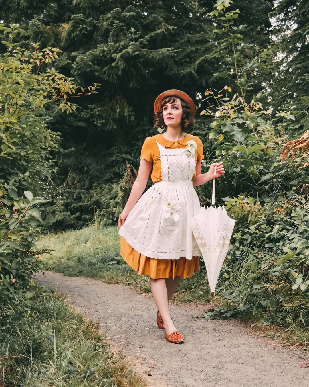 a fulllength photograph of a young woman strolling down a forest path. she is dressed in vintageinspired attire: a mustard yellow shortsleeved dress paired with a white, lacetrimmed apron adorned with small, embroidered daisies.  she wears a brown hat and brown shoes. a white lace umbrella is carried in her hand. the overall aesthetic is reminiscent of 1940s or 1950s fashion, with a whimsical and cottagecore feel. the setting is a lush, green forest with dappled sunlight filtering through the trees.  the woman appears serene and confident as she walks along the path.