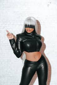 a woman confidently poses in a storm (xmen) inspired costume. she wears a black leather jacket and matching pants,  a silver wig, and a black blindfold. the image is taken indoors against a white brick wall, illuminated by bright artificial light. her pose and attire convey strength and power.