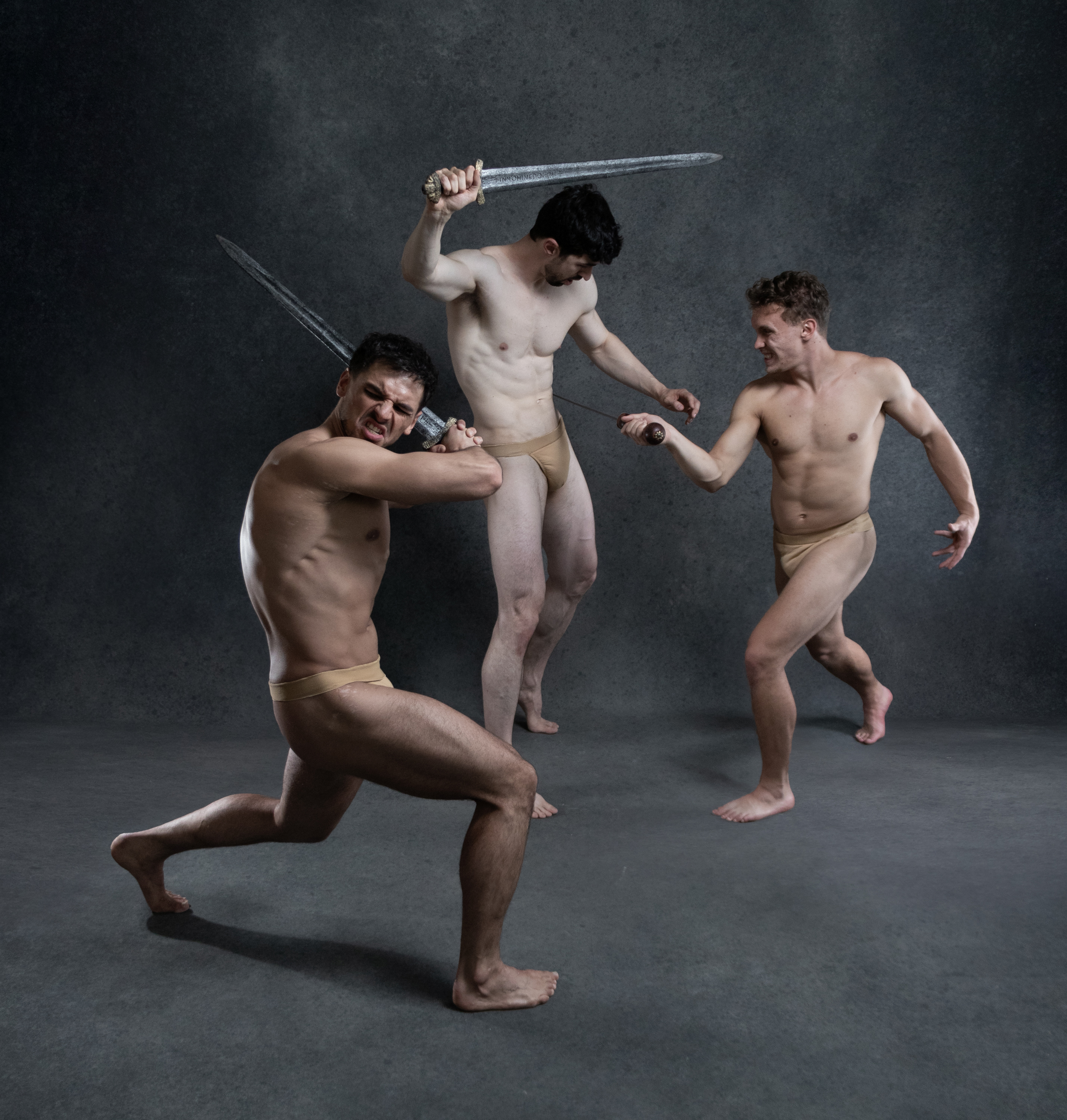 three muscular men in loincloths are engaged in a fierce sword fight against a dark studio backdrop. the scene is dramatically lit, highlighting the men's physiques and the tension of the battle. one man on the left defends himself with a raised sword, while another on the right prepares to strike. a third man in the center seems to be disarmed, but his determined expression suggests he's not out of the fight yet. the overall atmosphere evokes a sense of historical or mythological combat.