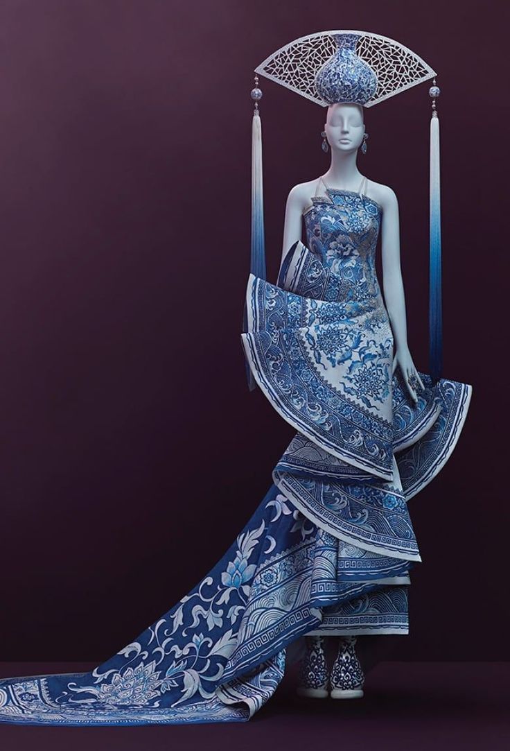 a fashion model is wearing a stunning blue and white dress inspired by chinese porcelain. the dress features an intricate pattern of flowers and geometric designs, and the headdress is also made of porcelain. the model is posing against a dark purple backdrop. the dress is elegant and luxurious, and the model is looking beautiful and creative. the intricate details on the dress make it look like a work of art.