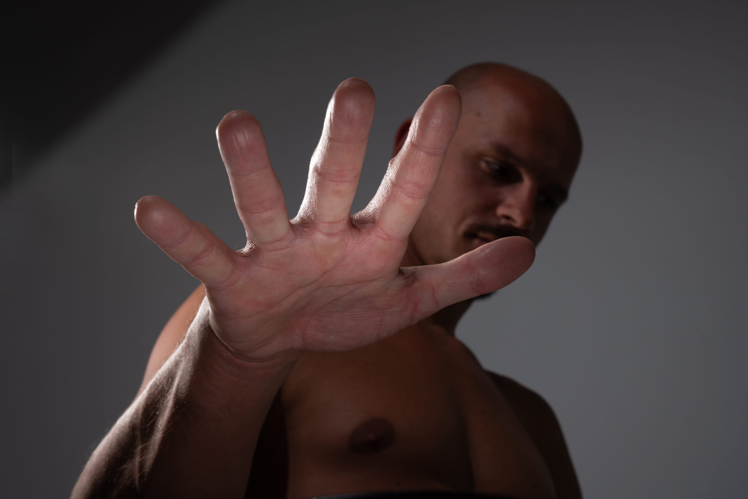 a shirtless man is holding up his hand in a "stop" gesture. the image is taken from a low angle, looking up at the man, making the hand the focal point of the image. the lighting is artificial and creates a dramatic contrast on his hand and body.