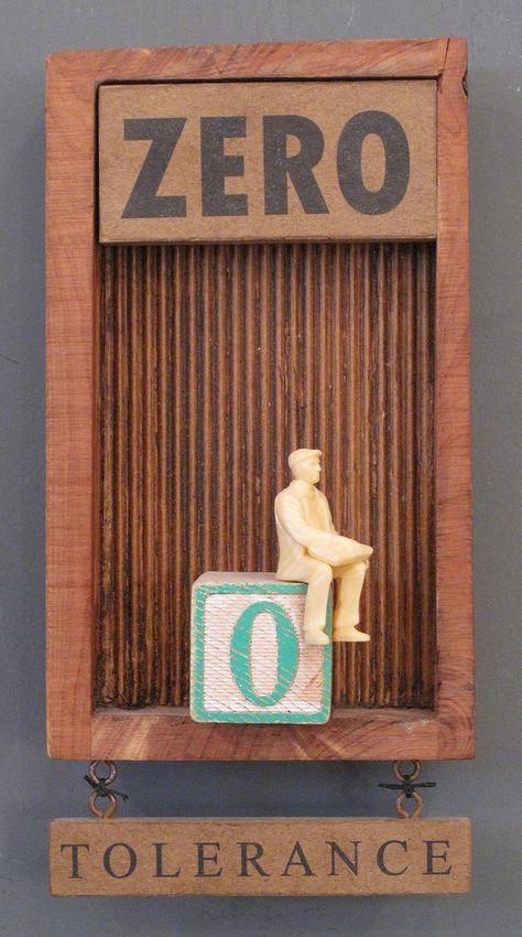 a closeup shot of an art piece with the words "zero" on the top, followed by a corrugated wooden section, with a small figurine sitting on a wooden block with the number "0" printed on it. below, a wooden slab hangs with the word "tolerance" written on it. the whole piece is framed with wood.