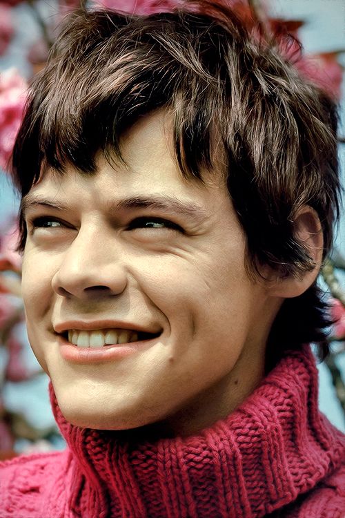 a closeup portrait of a young man, smiling brightly with a charmingly wide smile. he's wearing a red turtleneck sweater. his hair is brown and styled with a messy look. the setting appears to be outdoors, with pink flowers visible in the background. the overall mood of the image is happy and cheerful, capturing the man's natural charisma and positivity.
