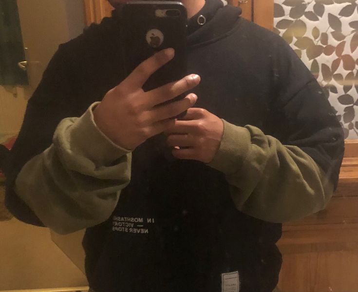 a person wearing a black hoodie with green sleeves is holding a phone to take a selfie in a mirror.