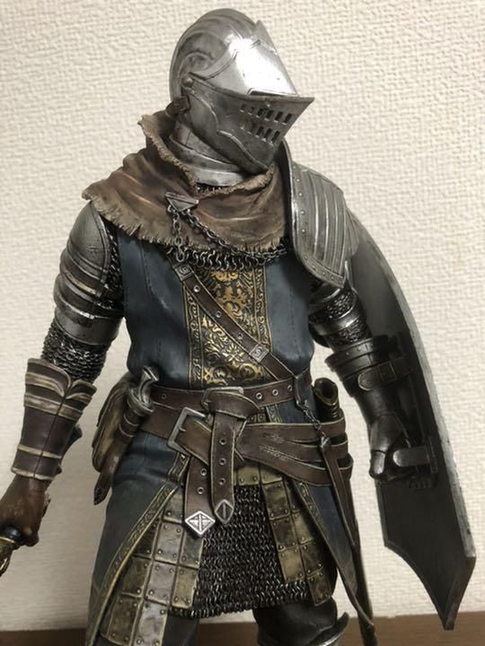 the image shows a highly detailed statue of a knight in full plate armor, reminiscent of the dark souls video game series. the knight is posed with one hand resting on a large shield and the other holding the hilt of a sword. the armor is intricately sculpted and painted, with a mix of silver, bronze, and leather textures. the figure stands against a plain white wall, highlighting the craftsmanship and details of the statue.