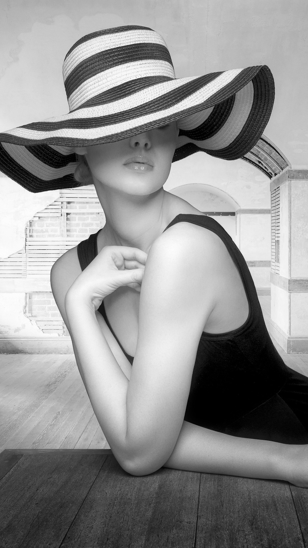 a black and white image of a woman in a striped sunhat and black tank top, sitting on a wooden floor with her arms crossed, with a light and shadowed background. the woman is looking directly at the camera.