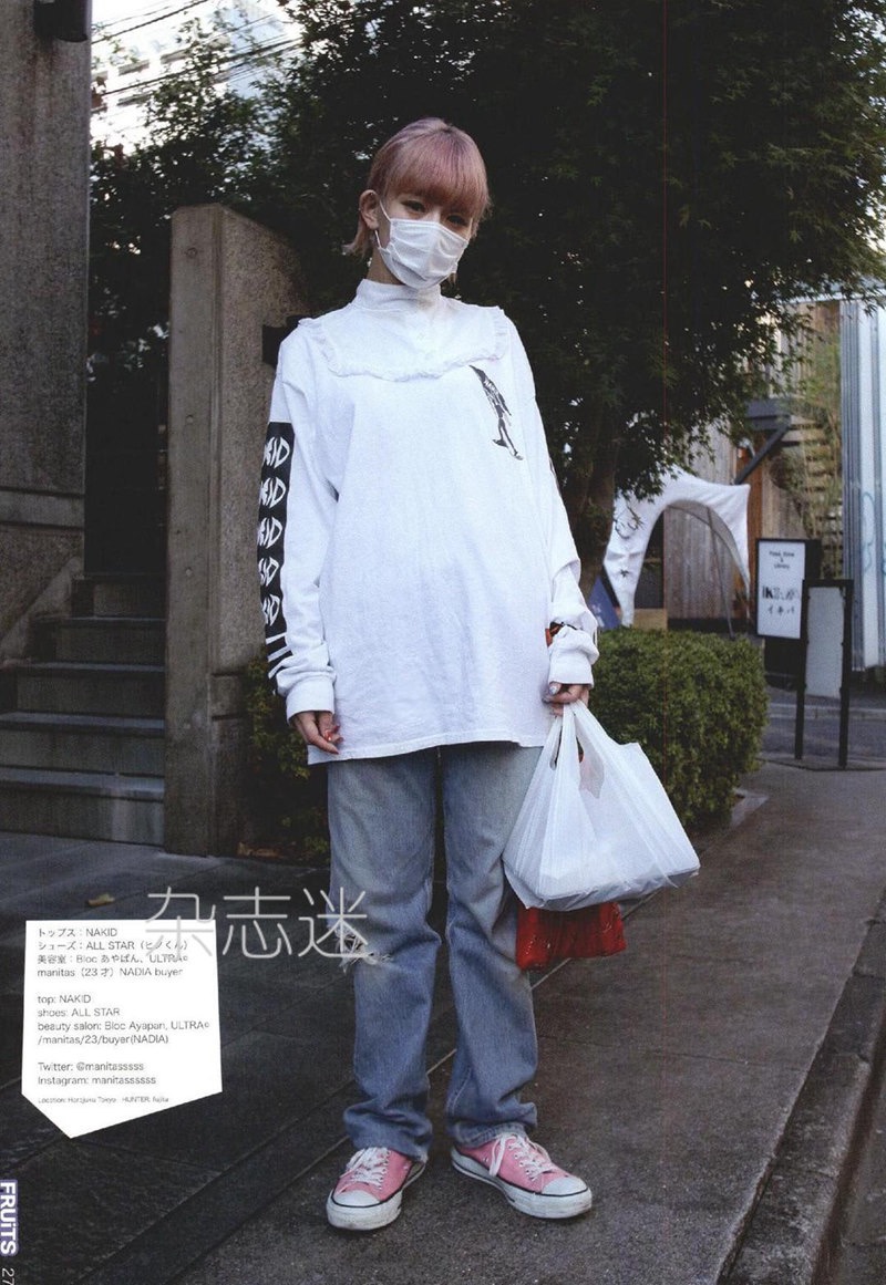 a young woman with pink hair and a face mask poses on a city street for a japanese fashion magazine. she wears an oversized white longsleeved shirt, loosefitting jeans, and pink converse sneakers. she carries a white shopping bag in her right hand. the image has a slightly desaturated and grainy aesthetic common in street photography.