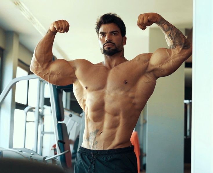 a muscular man is flexing his biceps and triceps while posing in a gym. he's wearing black athletic shorts and has a defined sixpack.  he has a number of tattoos on his arms. the background is a blurred shot of a gym.