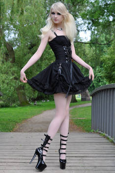 a young blonde woman in a black gothic lolita style dress poses on a wooden bridge in a park. she is wearing black platform boots with buckles and her dress has a corsetstyle bodice and a layered skirt. the setting is outdoors in a park with lush green trees. the photo is professionally taken, possibly for a fashion shoot.