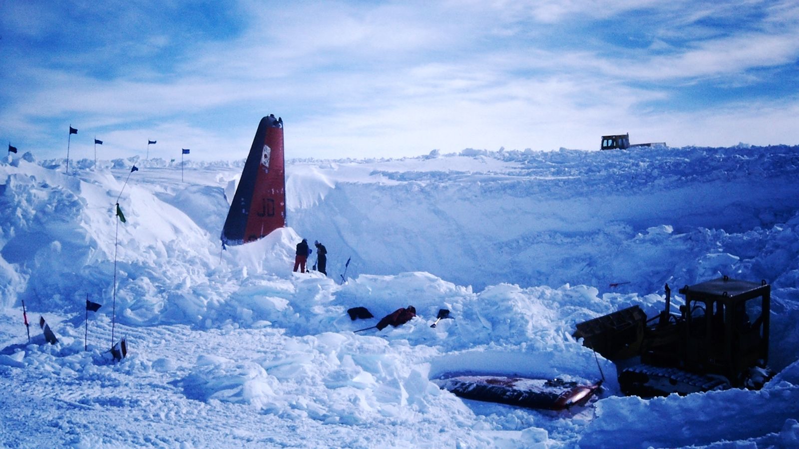 a snowcovered landscape with a partially buried plane wreckage. the plane is a red and white aircraft, with the tail section sticking out of the snow. there are several people standing around the wreckage, and a bulldozer is working in the background. the scene is very remote and desolate, with mountains in the distance. it is likely that this is a rescue operation in progress.