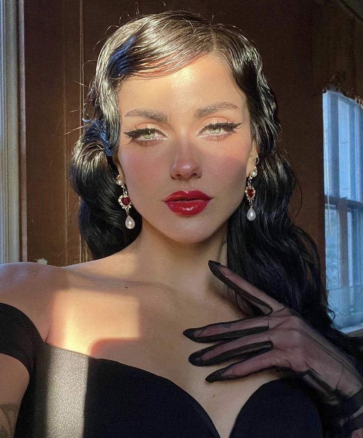 the image shows a woman with a vintageinspired look. she is wearing a black dress with a sweetheart neckline and black lace gloves. she has dark hair styled in a retro wave, with a side part and soft curls. her makeup features winged eyeliner, red lipstick, and rosy cheeks. she is wearing dangling earrings with red stones and pearls. the woman is lit by soft, natural light coming from a window, creating a warm and inviting atmosphere.