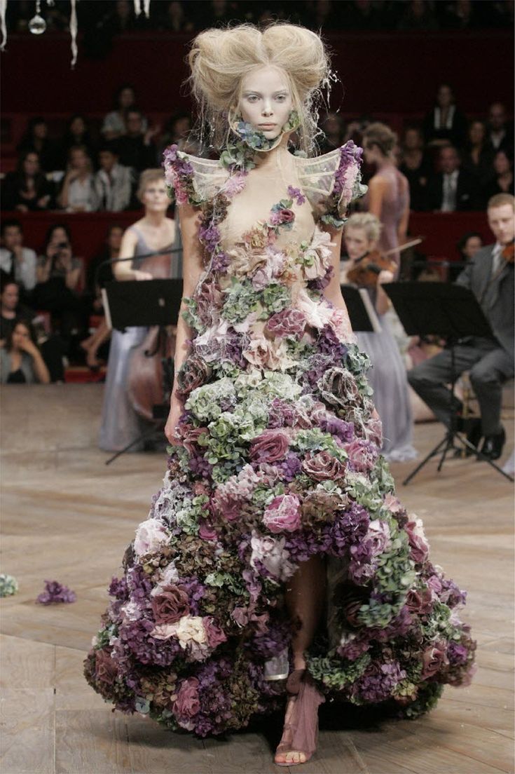 a model walks the runway in a stunning floral gown. the dress is made entirely of flowers, with layers of petals and leaves cascading down to the floor. the model's hair is styled in a dramatic updo, and she wears minimal makeup, allowing the dress to take center stage. the backdrop of the runway features a crowd of spectators watching the show. the dress is a showstopper, showcasing the creativity and artistry of the designer.