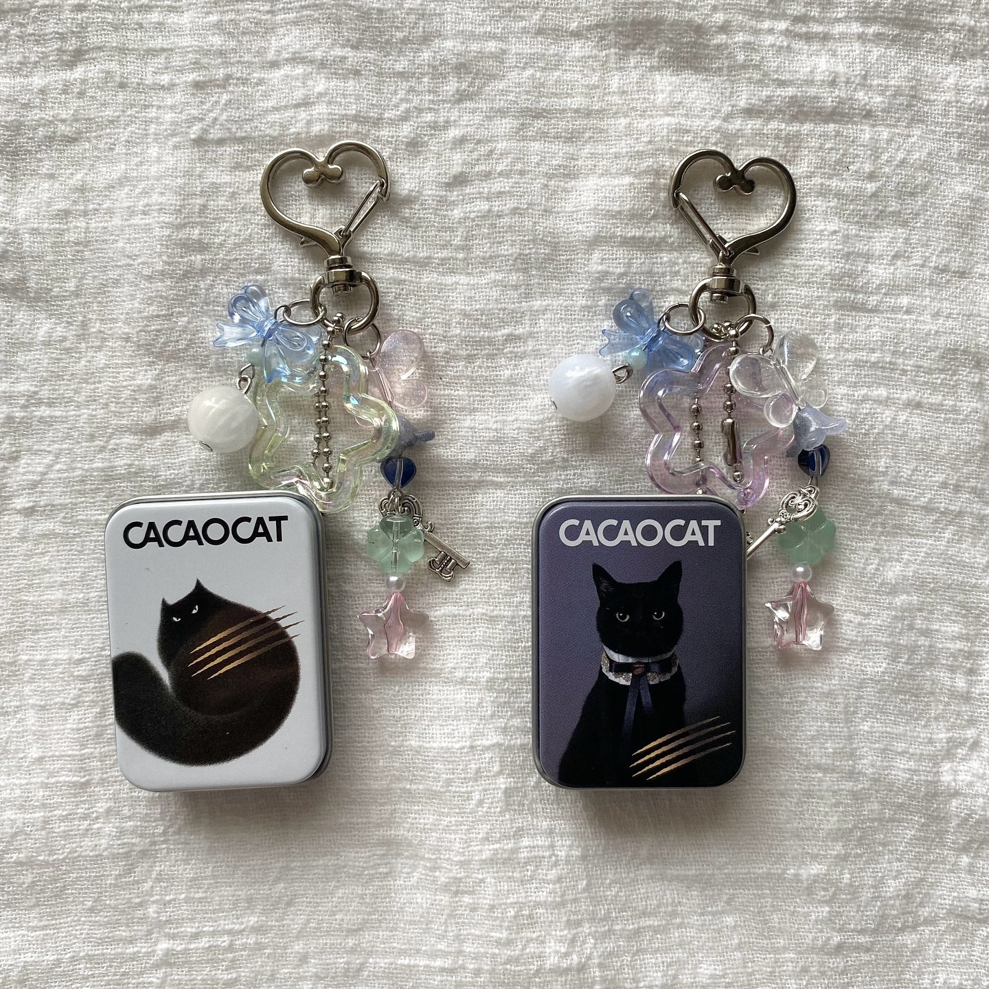 two cacao cat keychain with heartshaped clips and trinkets lie on a white textured background. the keychains are each attached to a small metal tin. each tin has a different image of a black cat on it. beads, charms, and other trinkets hang from the clips of the keychains. the overall aesthetic is pastel and whimsical.