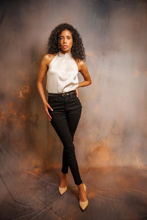 a fullbody studio shot of a confident woman with curly hair. she is wearing a creamcolored silk halter top, black skinny jeans, and nude high heels.  her pose is relaxed yet elegant, with her hand resting on her hip. the background is a textured, neutral tone, which helps her outfit stand out. the overall mood is sophisticated and stylish.