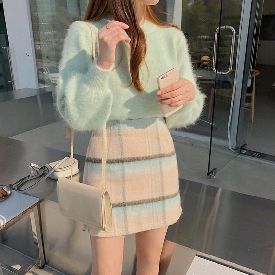 a woman is standing outdoors, holding a phone, wearing a mint green sweater and a plaid skirt. she is also wearing a light beige handbag. the outfit is in pastel colors. the woman is posing for the photo.