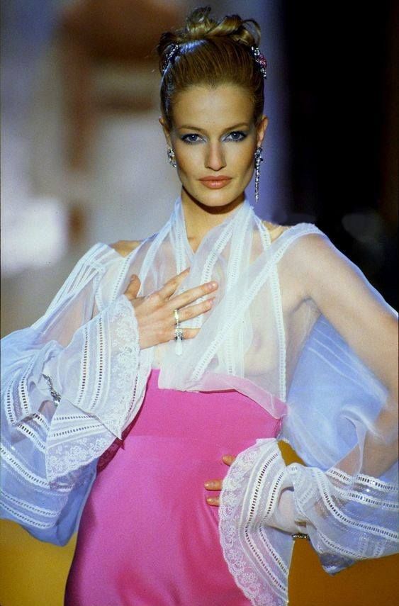 a woman is posing in a fashion show. she is wearing a pink dress with a sheer, white, longsleeved top with lace detailing. she has her hair up and is wearing earrings and other jewelry. she is on a runway or catwalk.