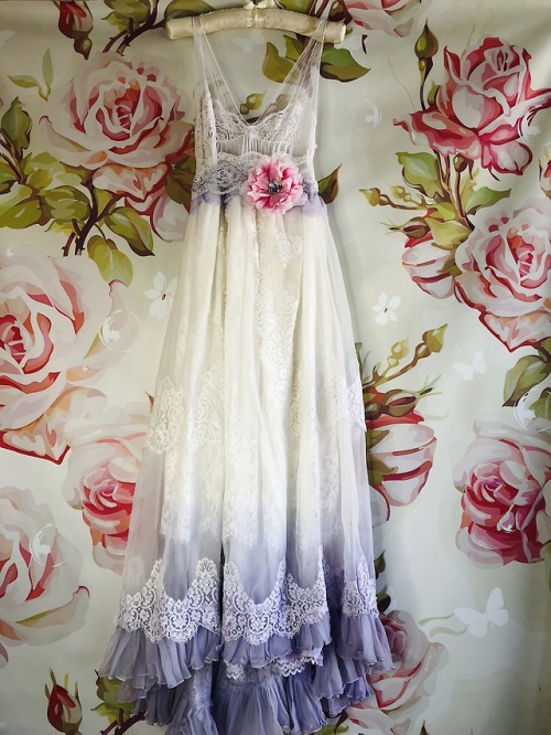 a long white and purple ombre wedding dress on a hanger. the dress is made of lace and has a floral sash. the background is a floral print.