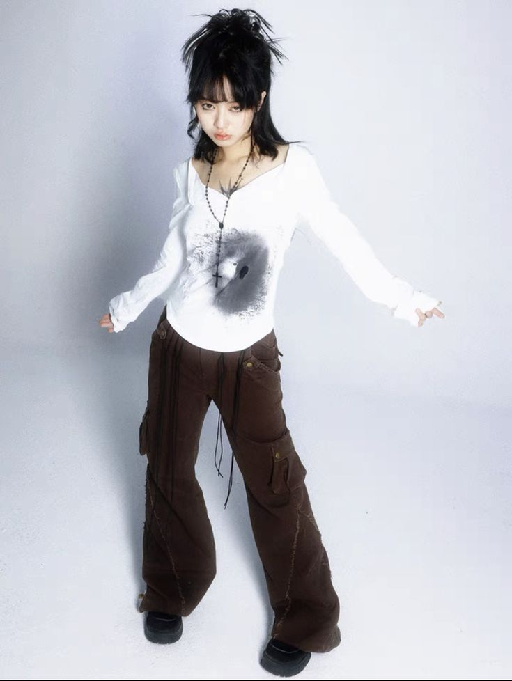 the image shows a young woman modeling an outfit. she has dark hair styled in a slightly messy, layered look. she's wearing a white longsleeved shirt with a dark graphic design on the front. the shirt has a slight vneck and slightly puffed sleeves.  over the shirt, she wears a delicate black cross necklace. her pants are wideleg, dark brown cargo pants with a distressed or worn look and visible seams. her shoes are dark, possibly black boots or shoes.  the overall style is a blend of grunge, alternative and possibly elements of 90s or y2k fashion. the background is plain white, focusing attention on the model and her clothing. the image appears to be a professional studio shot.