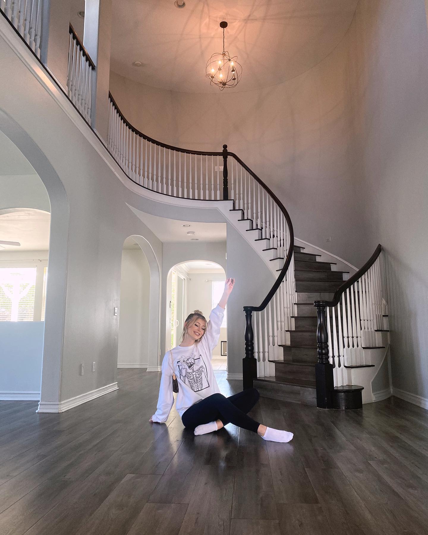 a woman sits crosslegged on the hardwood floor of a large, welllit home, raising one arm in the air. behind her are stairs with a dark wood railing and white balusters. the house has arched doorways and high ceilings, with a chandelier hanging from the ceiling.
