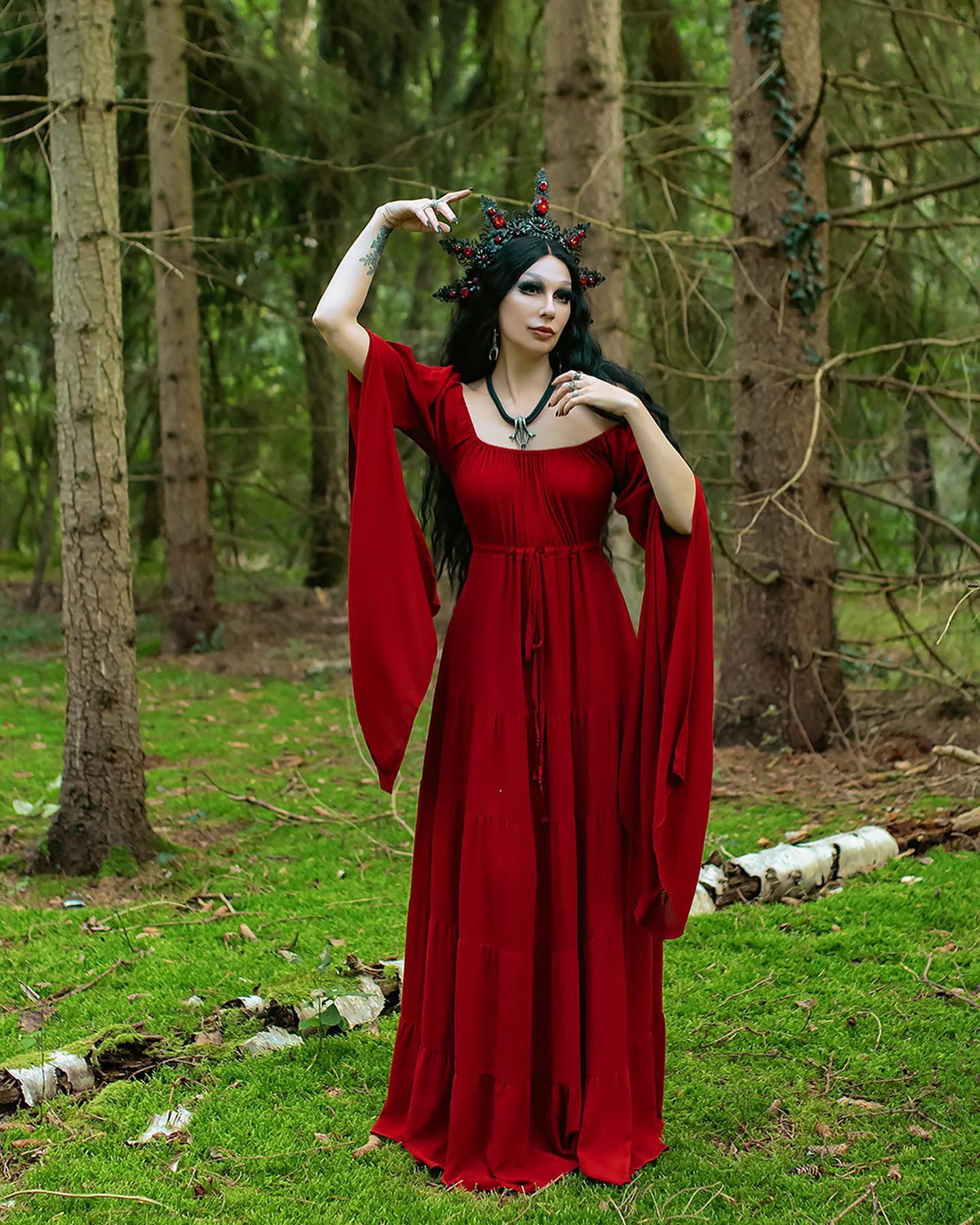 a woman in a long, flowing red medievalstyle dress stands in a forest. she wears a dark, ornate crown adorned with red gems and a necklace.  her long, dark hair cascades down her shoulders. the dress has wide, bellshaped sleeves, and the overall aesthetic is dark, gothic, or fantasyinspired. the setting is a lush green forest with mossy ground and tall trees. the scene is evocative of a dark fantasy or fairytale setting.  the model appears elegant and mysterious.