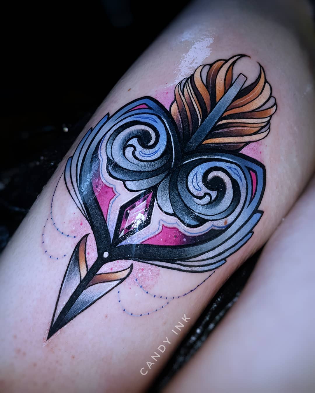 this is an image of a neo traditional tattoo of an arrow piercing a heart. the heart has decorative swirls on it and a pink and purple galaxy design in the background. the arrow's fletching is made of feathers done in shades of yellow and orange. there are strings of beads hanging from the bottom of the heart. the tattoo is on someone's inner thigh, just above the knee. the tattoo artist's watermark, "candy ink" is written in all caps on the person's thigh below the tattoo.