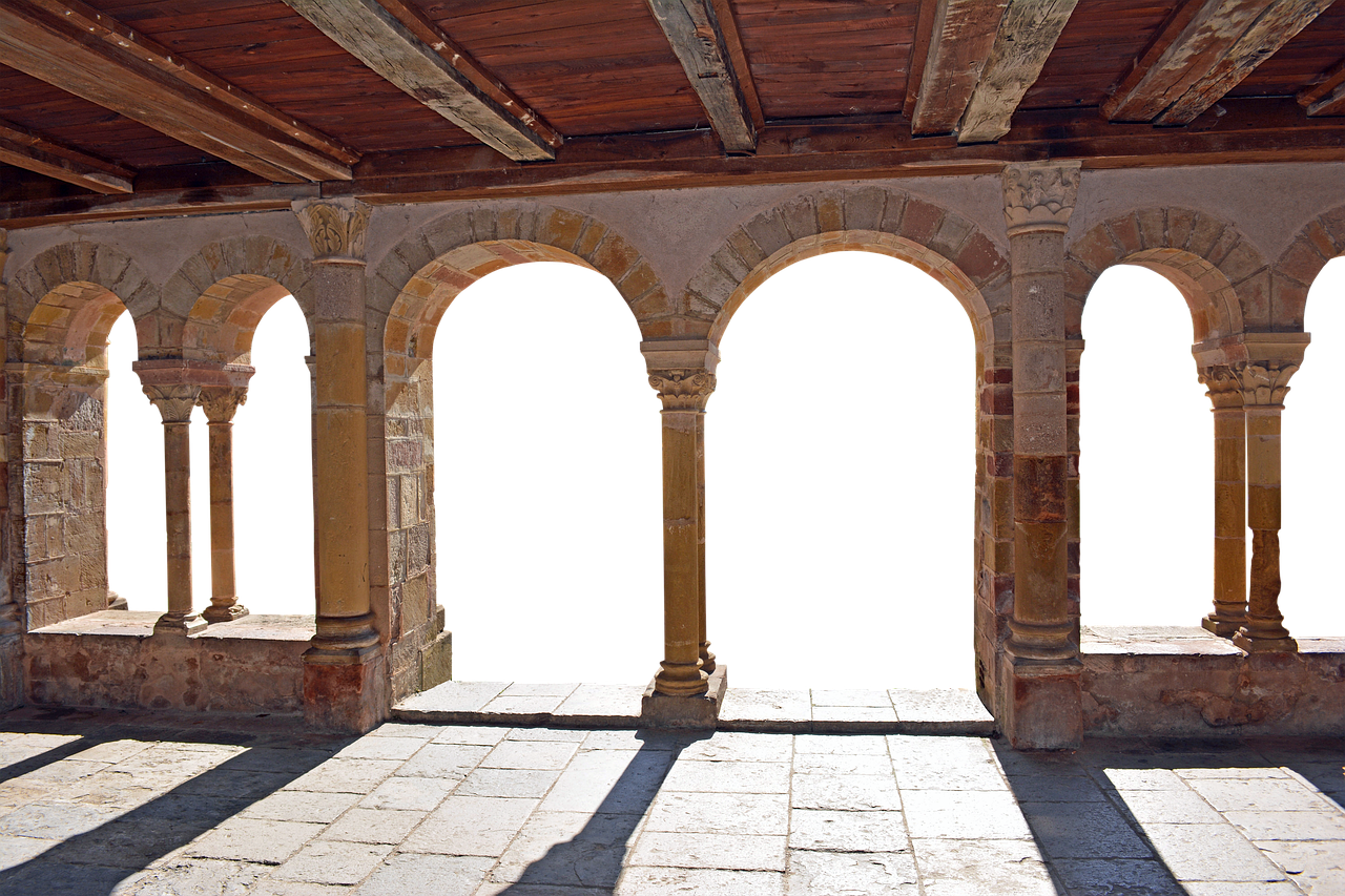 the image depicts a cloister with arches and columns, creating a hallway. the structure is made of stone, and shadows cast by the sunlight add depth to the scene. the building's interior is visible, showcasing the architectural details of the arches and columns.