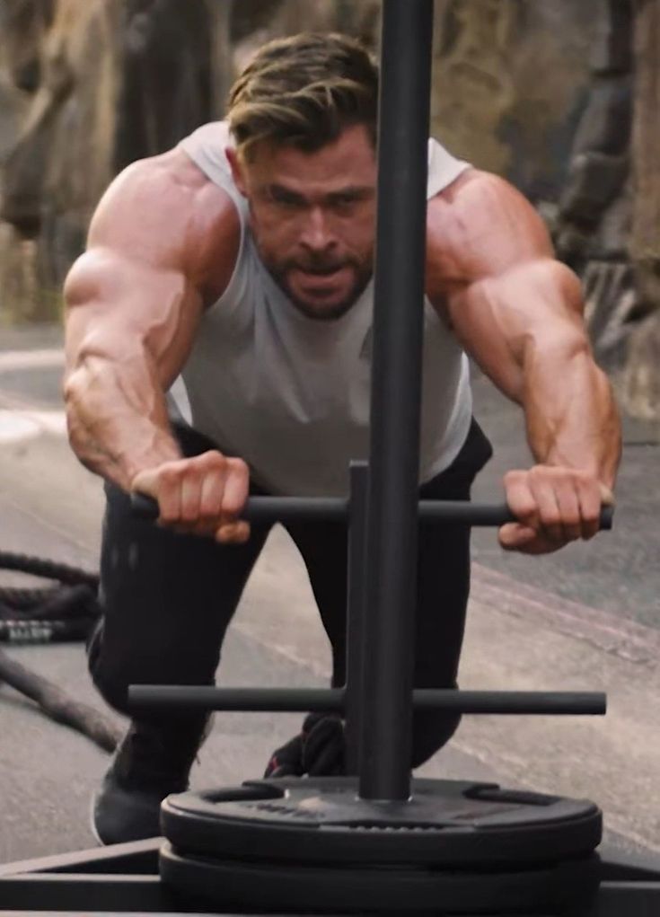 chris hemsworth is seen in a closeup portrait, engaged in an intense workout session. he's utilizing a heavyduty weightlifting contraption with a weight plate attached. his muscular physique is evident as he pushes the weight with sheer strength and determination, showcasing his dedication to fitness and his role as thor.