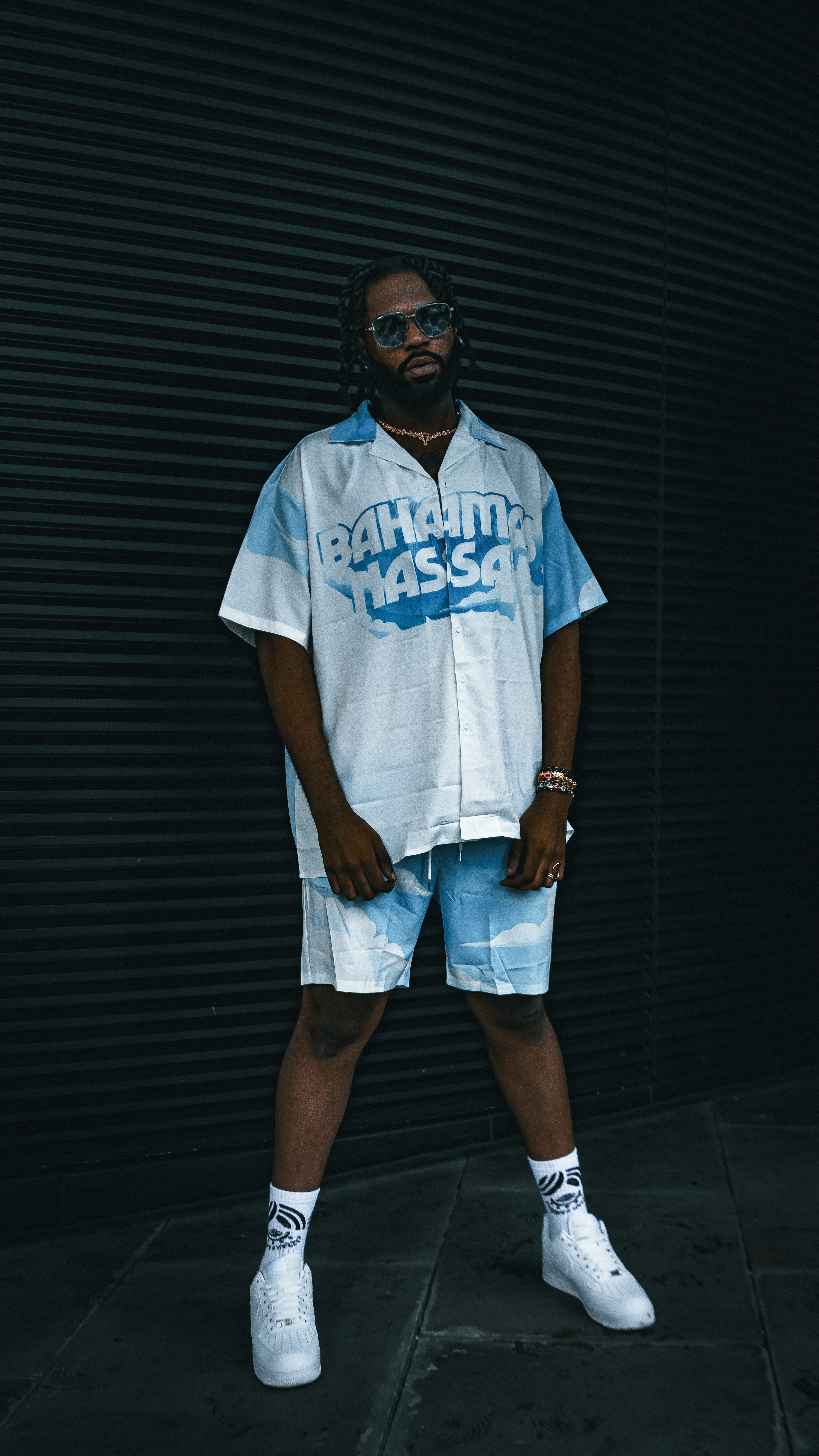 a fullbody shot of a man standing against a black wall. he is wearing a white and light blue printed shirt and matching shorts. his outfit is completed with white sneakers and dark sunglasses. the man has a beard and dark hair and is looking towards the camera.